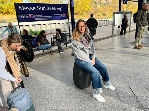 Eine Frau mit Brille, grauer Jacke und Jeans sitzt an der Berliner S-Bahn-Haltestelle „Messe Süd (Eichkamp)“ auf einem großen, schwarzen, kissenartigen Gegenstand, der als Sitzgelegenheit dient.