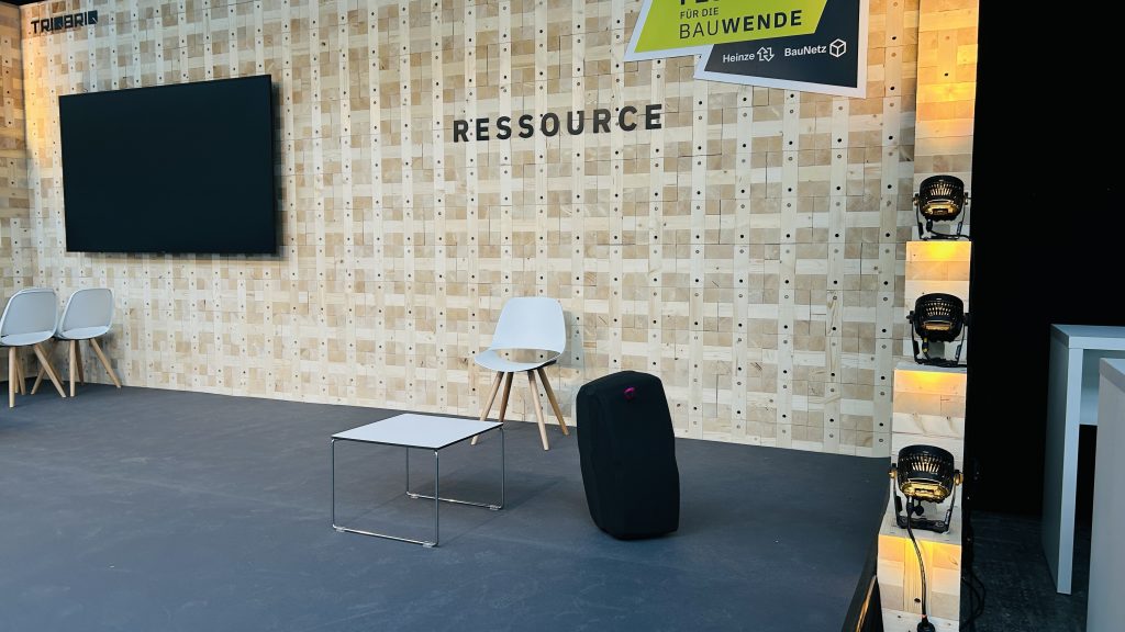 Ein moderner Messestand mit einer Holzwand aus quadratischen Holzblöcken, die das Wort „RESSOURCE“ zeigt, einem Bildschirm, einer weißen Bestuhlung und dem schwarzen, kissenartigen Sitzmöbel.