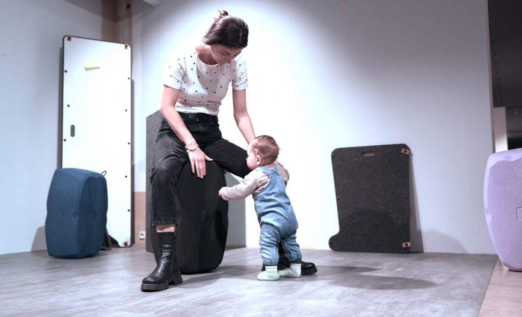 Eine junge Frau sitzt auf einem großen, runden, dunkelgrauen Möbelstück oder Designobjekt. Sie trägt ein weißes T-Shirt mit dunklen Punkten, schwarze Jeans und dunkle, klobige Stiefel. Vor ihr steht ein Kleinkind in blauer Jeans-Latzhose und hellgrünen Socken, das sich an dem Objekt festhält und zur Frau aufblickt. Der Raum wirkt wie ein modernes Studio oder Ausstellungsraum mit hellem Boden und weißen Wänden, im Hintergrund stehen weitere abstrakte, gepolsterte Möbelstücke.