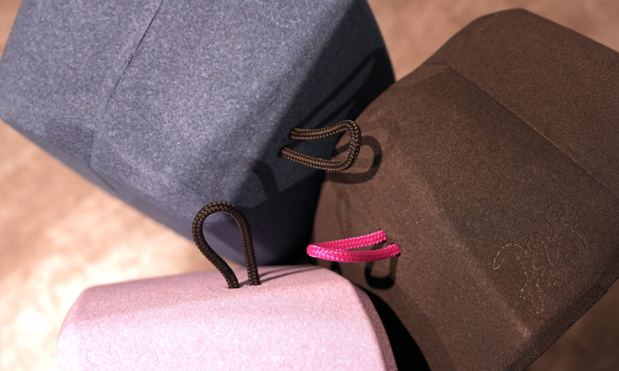 Drei würfelförmige, gepolsterte Hocker-Blöcke, aus einer leichten Aufsicht aufgenommen, die sich in der Mitte des Bildes berühren. Die Farben sind Dunkelblau (Black/Blueberry), Dunkelbraun und Hellrosa (Pale Lavender). Jeder Block hat eine farblich abgesetzte Stoffschlaufe zum Tragen: zwei Blöcke haben dunkelbraune/schwarze Schlaufen, der rosa Block hat eine leuchtend pinke Schlaufe. Die Blöcke zeigen eine subtile, Ton-in-Ton-Musterung.