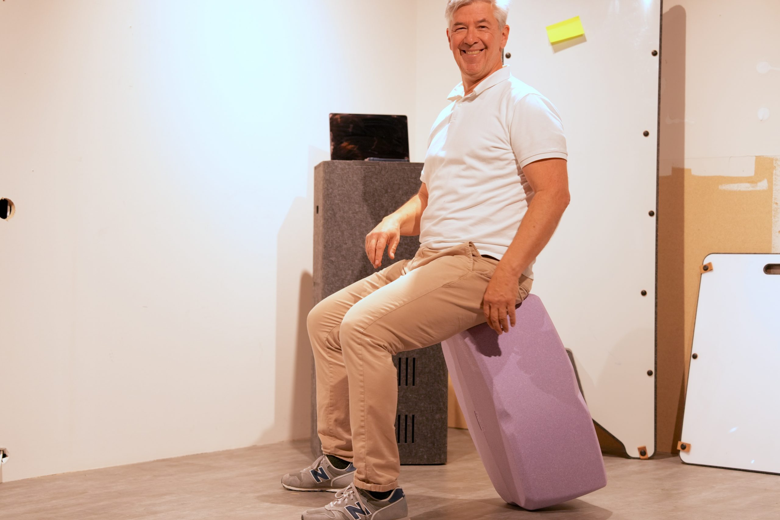 Ein lächelnder, älterer Mann sitzt aufrecht auf einem hohen, fliederfarbenen Keilkissen, das als ergonomischer Hocker dient.
