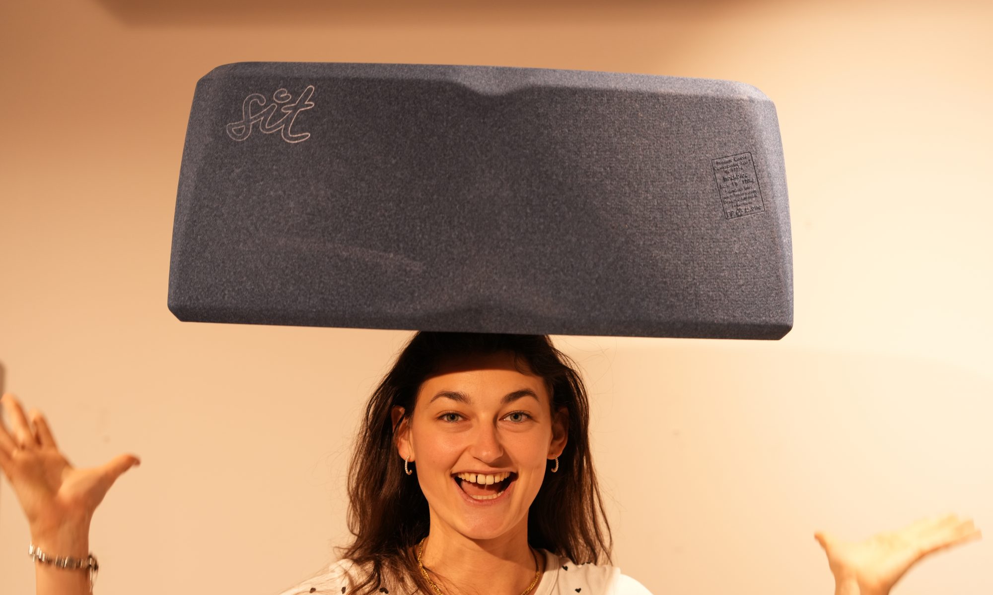 Eine lächelnde junge Frau balanciert ein großes, blaues, keilförmiges Sitzkissen mit der Aufschrift „sit“ auf ihrem Kopf.