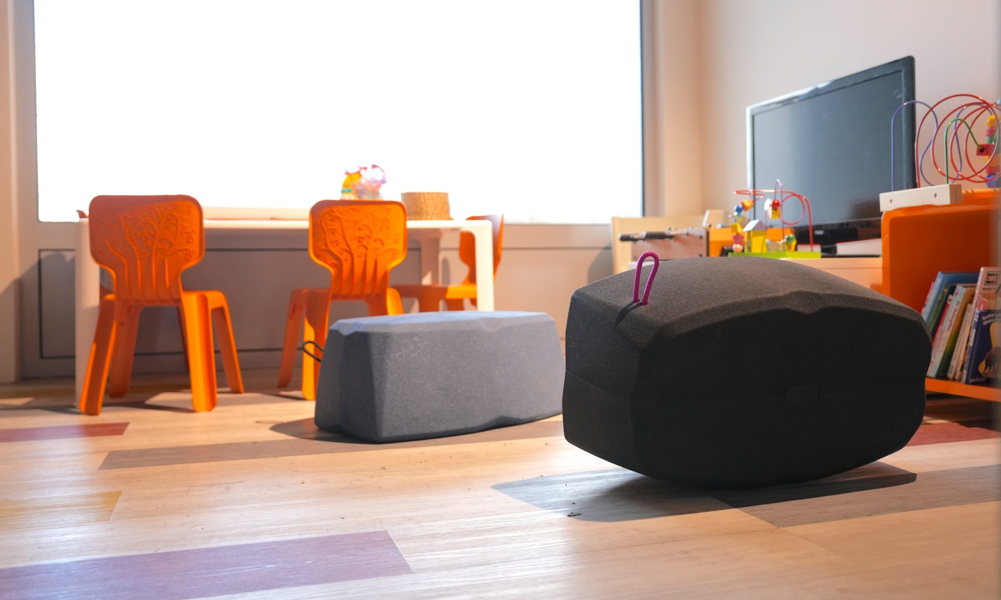 Zwei flache, gepolsterte Hocker-Blöcke liegen im Vordergrund eines Spielzimmers oder Kinderbereichs mit Holzboden. Einer ist dunkelgrau (Black/Blueberry) mit einer leuchtend pinken Trageschlaufe, der andere ist einfarbig grau. Im Hintergrund, vor einem großen Fenster, stehen ein niedriger, weißer Kindertisch und zwei leuchtend orangefarbene Kinderstühle. Rechts befindet sich ein Fernseher und Spielzeug, was auf eine kinderfreundliche Nutzung des Raumes hinweist.