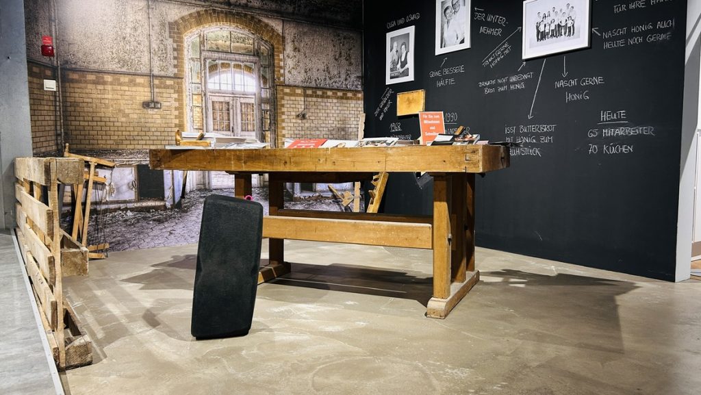 Ein Ausstellungsraum mit einem rustikalen Holzarbeitstisch, einer Fototapete mit einer alten Fabrikhalle, einer Tafelwand mit handschriftlicher Chronik und dem schwarzen, kissenartigen Sitzmöbel.