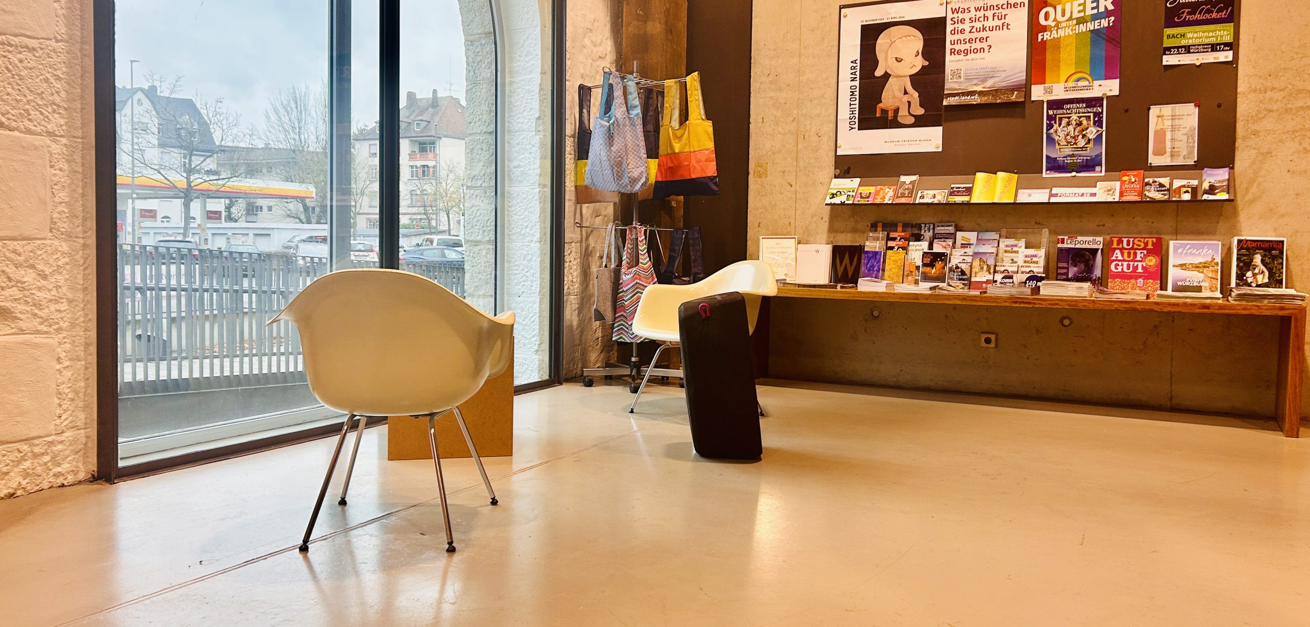 Eine junge Frau sitzt in einem großen, leeren Veranstaltungsraum auf einem schwarzen, kissenartigen Sitzobjekt, hält ein rotes Widerstandsband und lächelt in die Kamera. Der Raum ist dramatisch lila und blau beleuchtet.