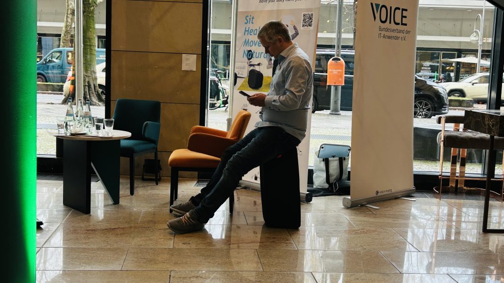 Ein Mann sitzt in einem Hotel- oder Messefoyer auf einem schwarzen, kissenartigen Sitzobjekt, beugt sich nach vorne und schaut auf sein Smartphone. Im Hintergrund sind Ausstellungsbanner und die belebte Stadtstraße sichtbar.