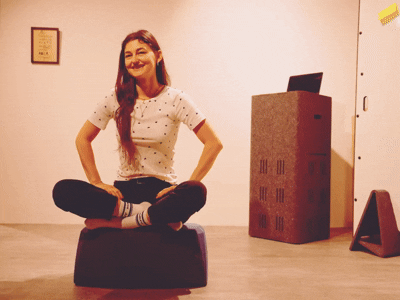 Ein kurzes GIF zeigt eine lächelnde junge Frau, die im Schneidersitz auf einem ergonomischen, blauen Sitzkissen am Boden sitzt.