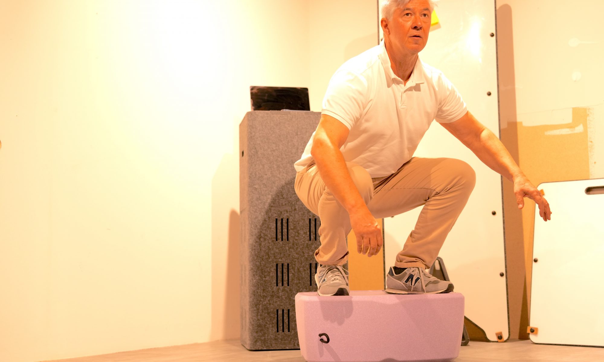 Ein älterer Mann führt eine tiefe Kniebeuge stehend auf einem stabilen, fliederfarbenen Sitzkissen aus, um dessen Belastbarkeit