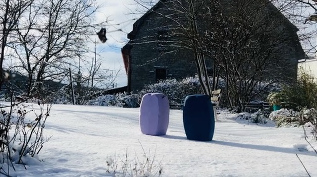 Zwei moderne, tonnenförmige Sitzmöbel in Lila und Dunkelblau stehen auf einer schneebedeckten Wiese vor einem rustikalen Steinhaus und einer Gartenbank im Winter.
