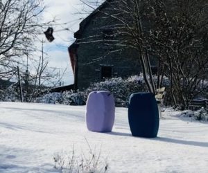 Zwei moderne, tonnenförmige Sitzmöbel in Lila und Dunkelblau stehen auf einer schneebedeckten Wiese vor einem rustikalen Steinhaus und einer Gartenbank im Winter.