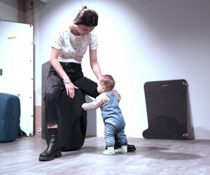 Eine junge Frau sitzt auf einem großen, runden, dunkelgrauen Möbelstück oder Designobjekt. Sie trägt ein weißes T-Shirt mit dunklen Punkten, schwarze Jeans und dunkle, klobige Stiefel. Vor ihr steht ein Kleinkind in blauer Jeans-Latzhose und hellgrünen Socken, das sich an dem Objekt festhält und zur Frau aufblickt. Der Raum wirkt wie ein modernes Studio oder Ausstellungsraum mit hellem Boden und weißen Wänden, im Hintergrund stehen weitere abstrakte, gepolsterte Möbelstücke.